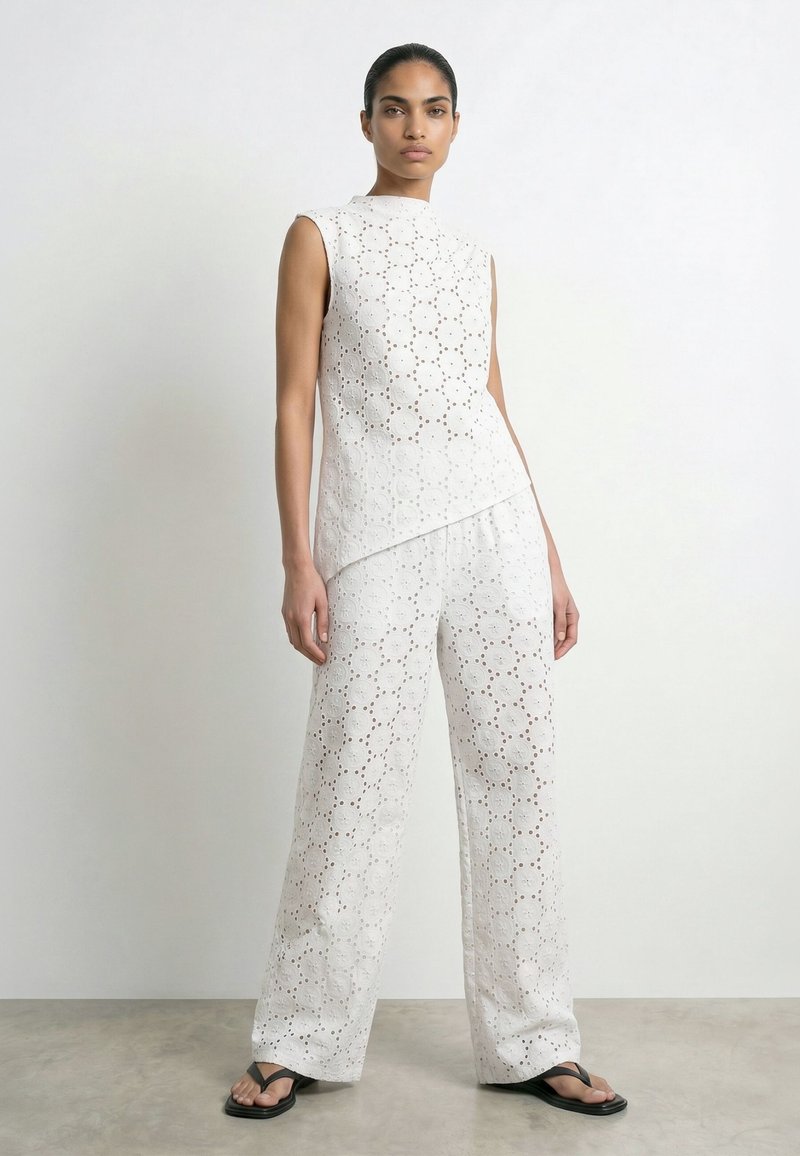 Femme debout portant un haut blanc sans manches et un pantalon large assorti avec broderie œillets, associé à des sandales noires, fond blanc.