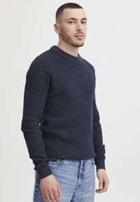 Sötétkék kötött pulóver kerek nyakkivágással, bordás mandzsettákkal és struktúrált mintázattal, világos farmernadrággal párosítva.