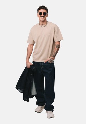 Mężczyzna z tatuażami i okularami przeciwsłonecznymi, ubrany w beżowy t-shirt, ciemne dżinsy, białe trampki, trzymający czarną kurtkę na jednolitym tle.