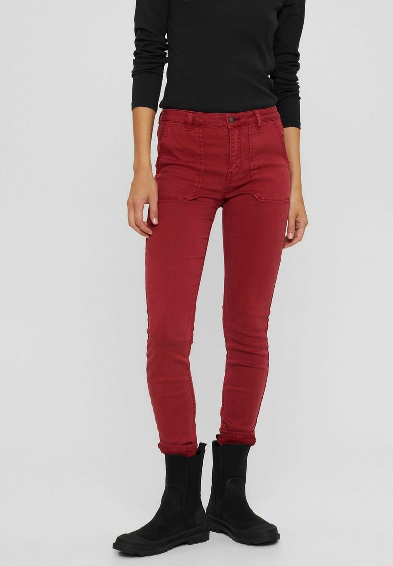 Jean skinny en coton rouge avec poches avant et passants de ceinture, accompagnés de bottines noires. Le tissu a une texture lisse et un design ajusté.