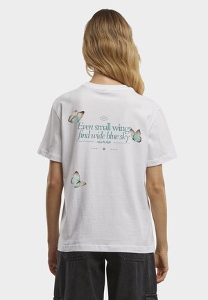 Kobieta z długimi włosami w białej koszulce z turkusowymi motylami i napisem „Nawet małe skrzydła znajdą szerokie, niebieskie niebo” na plecach.