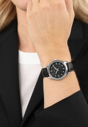 Schwarze Armbanduhr mit einem silbernen Metallgehäuse, schwarzem Zifferblatt und silbernen Stundenmarkierungen. Verfügt über ein schwarzes Lederarmband und schlichte Designdetails.