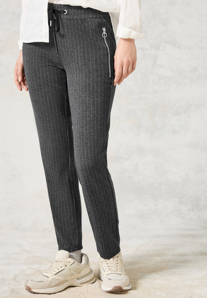 Pantalons gris à rayures, fabriqués en tissu doux, avec une taille élastique confortable et une poche zippée sur le côté, associés à des baskets neutres.