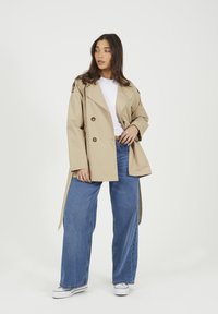 Beżowy podwójnie zapinany trencz o luźnym kroju, z dużymi guzikami, szerokimi rękawami i otworem z przodu, w połączeniu z jasnoniebieskimi spodniami szerokimi nogawkami.