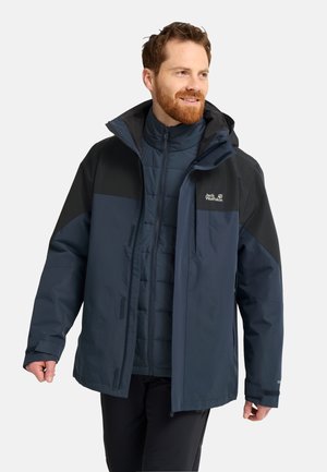 Mann trägt eine marineblaue und schwarze Outdoorjacke mit Kapuze, steht und lächelt, während er zur Seite schaut.