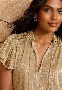 Gold shimmer top with ruffled sleeves, textured fabric, a V-neckline, and decorative stitching along the collar. Accented with earrings.