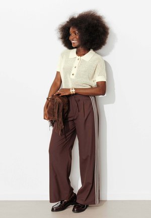 Femme avec une grande coiffure afro portant un polo en maille crème, un pantalon large marron avec des bandes latérales, tenant un sac en daim marron à franges, souriante, debout contre un mur blanc.