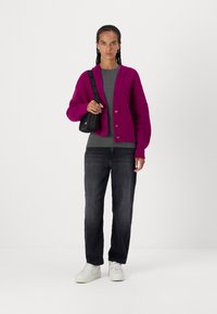 Cardigan lavorato a maglia fucsia con maniche a coste, top grigio, jeans scuri e sneakers bianche. Porta una piccola borsa a tracolla nera con tracolla con logo.