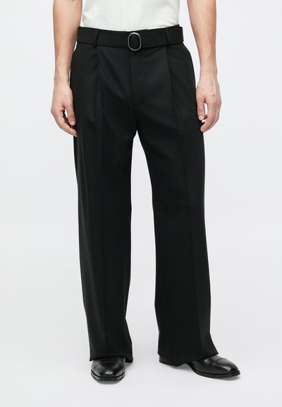 Juodos pritaikytos kelnės su juosmens diržu, kuriame yra apskritas metalinis dirželio sagtis, lygia priekiu ir plačiomis kojų kelnėmis. Lygios audinio tekstūros.