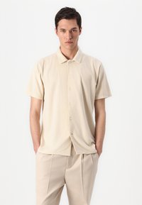 Μπεζ κοντομάνικο πουκάμισο με κουμπιά, κατασκευασμένο από μαλακό ύφασμα, με γιακά και ευθύ στρίφωμα. Τα μπροστινά κουμπιά σε ανοιχτό χρώμα συμπληρώνουν το σχέδιο.