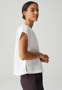 Weißes Kurzarmoberteil mit entspannter Passform, seitlichen Schlitzen und einer sanften, glatten Textur. Kombiniert mit dunklen Hosen, seitlich betrachtet.