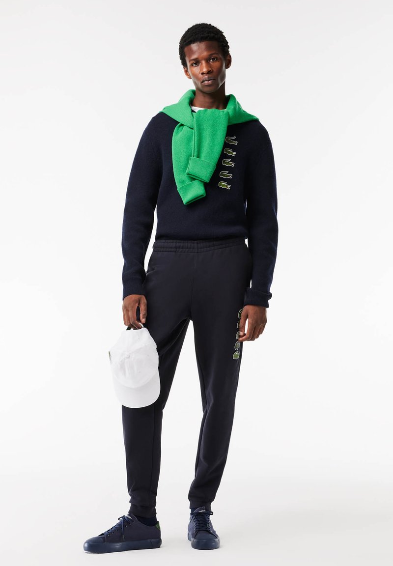 Pull marin bleu avec logo de grenouille verte, écharpe verte, jogging marin avec accents verts, casquette blanche tenue à la main, baskets bleu foncé.