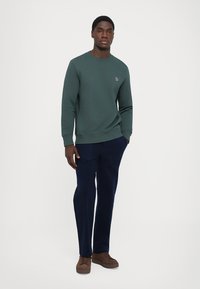 Hombre que lleva un sudadera verde oscuro, pantalones azul marino y zapatillas marrones, de pie con una mano en el bolsillo frente a un fondo blanco.
