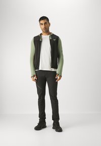 Zwarte en groene sportvest met capuchon, lange groene mouwen, gecombineerd met zwarte broek en donkere schoenen, met logo- en ritsdetails.