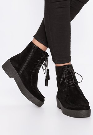 Black suede ankle boots with a chunky rubber sole, featuring laces and tassels, and a side zipper for easy wear.