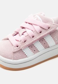 Pink suede sneaker with white stripes, pink laces, and gum sole close-up showing texture and stitching details.