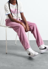 Lyserøde fløjlbukser, hvid t-shirt med blå kant og pink logo, hvide høj-ankel sneakers med sort swoosh, model siddende på stol.