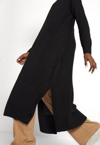 Schwarzes, geripptes Stricklangkleid mit seitlichen Schlitzen, kombiniert mit strukturierten beigen Weite-Hosen und weißen Turnschuhen. Fokus auf die Struktur des Stoffes.