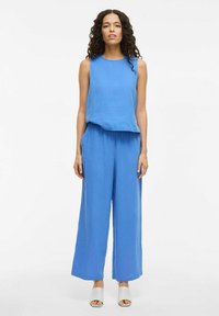 Blauw tweedelig outfit met een mouwloze top en wijde broek van een gestructureerde stof, voorzien van zijzakken. Gecombineerd met witte slip-on sandalen.