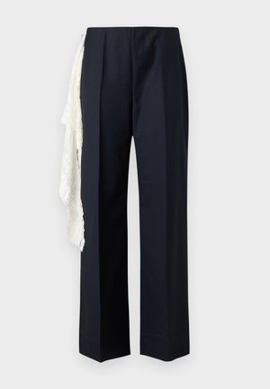 Žena s rovnými tmavými vlasmi stojí s rukami na bokoch, oblečená v čiernom tričku s dlhým rukávom, široké námornícke nohavice s bielym čipkovaným detailom a čierne podpätky.