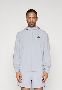 Ανοιχτό γκρι μπουφάν με φερμουάρ και κουκούλα, με μαύρο λογότυπο Adidas, ελαστικά μανίκια και αεριζόμενα σορτς από διχτυωτό ύφασμα. Ελαφρύ, αθλητικό σχέδιο.