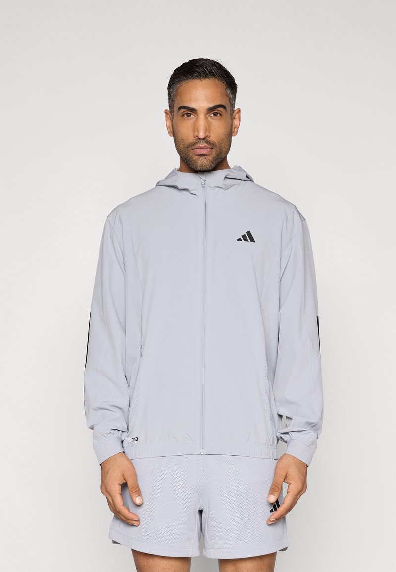 Ανοιχτό γκρι μπουφάν με φερμουάρ και κουκούλα, με μαύρο λογότυπο Adidas, ελαστικά μανίκια και αεριζόμενα σορτς από διχτυωτό ύφασμα. Ελαφρύ, αθλητικό σχέδιο.