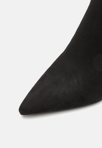 Chaussure en daim noir à bout pointu, présentant une texture lisse et un design épuré. Le talon est fin, ajoutant de l'élégance à la silhouette.