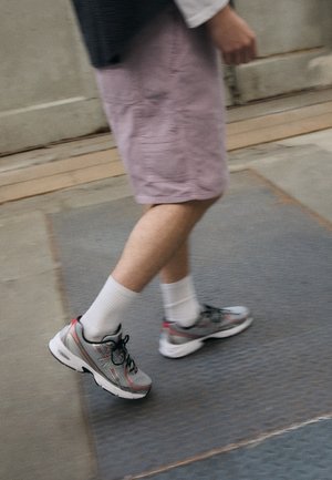 Person geht drinnen und trägt graue und rote Sneakers, weiße Socken und hellviolette Shorts auf einem gefliesten Boden.