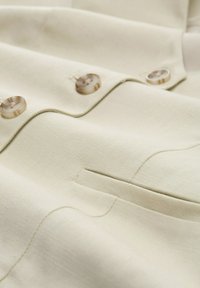 Helle beigefarbene Stoff mit strukturiertem Webmuster, mit zwei runden, hornartigen Knöpfen und einem kleinen Taschendetail.
