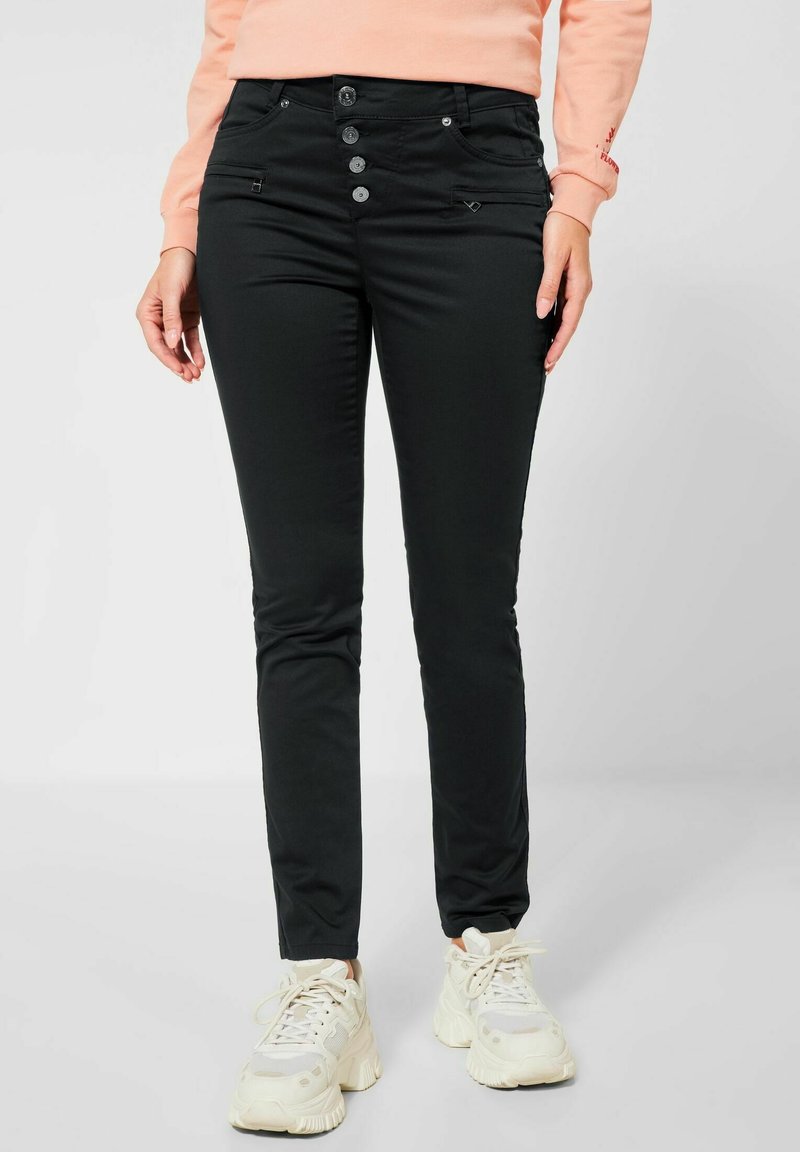 Femme portant un pantalon slim noir à boutons, un haut à manches longues pêche et des baskets blanches épaisses, debout devant un fond clair uni.