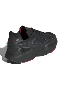 Scarpa sportiva nera con tomaia testurizzata, strisce viola e dettagli rossi. Presenta un colletto imbottito e una suola spessa con battistrada pronunciato.