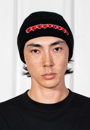 Młoda osoba z krótkimi ciemnymi włosami i piegami, ubrana w czarną czapkę z czerwonym napisem "PLEASURES" oraz czarną bluzę, o neutralnym wyrazie twarzy.