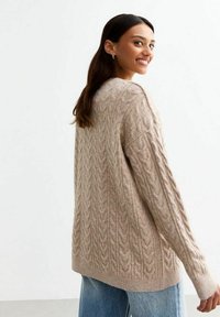 Suéter de punto de cable en color beige con mangas largas y un corte relajado, que presenta patrones texturizados y un cuello acanalado, combinado con jeans de color azul claro.