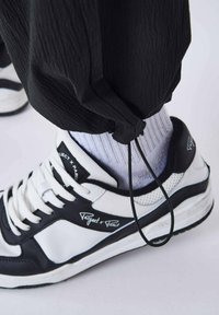 Hvide og sorte sneakers med tekstureret materiale, snørebåndsdesign, mesh-accenter og "Project x Paris" branding. Justerbar sort snøre-detajle.