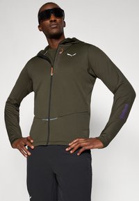 Αθλητικό μπουφάν σε ελιά με κουκούλα, φερμουάρ και οπές για τον αντίχειρα; διαθέτει αντιθέσεις σε μοβ αποχρώσεις και ανακλαστικές λεπτομέρειες.