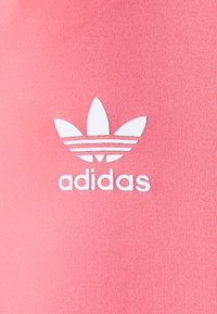 Tecido rosa com uma textura suave apresenta um logótipo da Adidas em branco, incluindo três riscas semelhantes a folhas acima do nome da marca em fonte negrito.