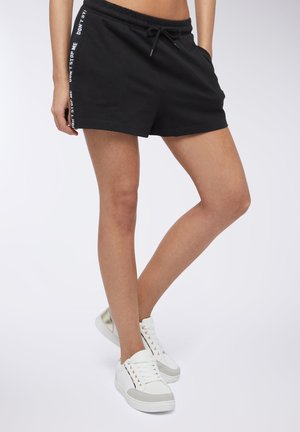 Shorts sportivi neri in cotone con vita elastica e coulisse, con testo bianco lungo il lato. Abbinati a sneakers bianche.