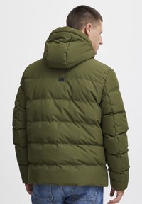 Jachetă verde matlasată cu glugă, având secțiuni cusute și un mic logo pe spate. Fabricată dintr-un material neted și durabil.