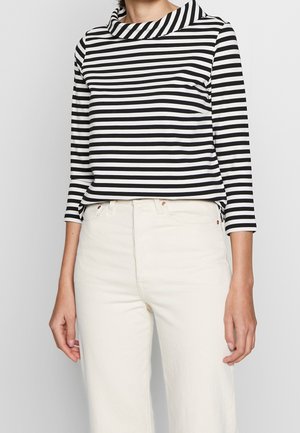 Person iført en sort og hvid vandret striped langærmet top med en rullekrave og lyse, højtaljede bukser.