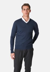 Maglione blu navy a scollo a V realizzato in tessuto lavorato a maglia, con maniche lunghe e polsini e orlo a coste. Indossato sopra una camicia bianca con colletto.