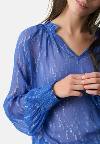 Transparente blaue Bluse mit V-Ausschnitt, gerafftem Halsauschnitt und Rüschensch cuffs. Mit silbernen Punktmustern für Textur und Leichtigkeit.