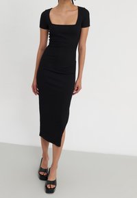 Kvinna i svart midiklänning med fyrkantig halsringning och korta ärmar, med asymmetrisk nederkant, som bär svarta plattformsskor med öppen tå, stående på en ljus golv.