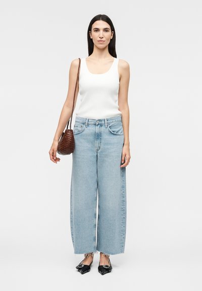 Γυναίκα φοράει λευκή αμάνικη μπλούζα, ανοιχτό μπλε τζιν με φαρδιά μπατζάκια, μαύρα μυτερά παπούτσια και κρατά καφέ πλεγμένη τσάντα ώμου.