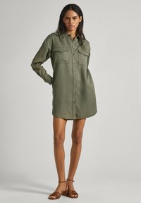 Abito camicia verde oliva realizzato in tessuto morbido, con chiusura a bottoni sul davanti, due tasche sul petto, maniche lunghe e orlo leggermente curvato.