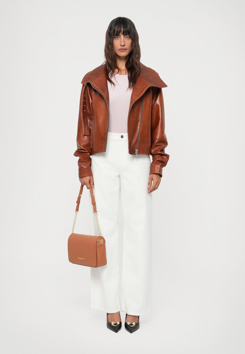 Veste en cuir marron avec un large col, haut rose clair, pantalon blanc taille haute, tenant un sac à main marron et portant des talons noirs avec des accents dorés.