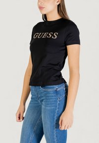 Camiseta negra de algodón con mangas cortas que presenta un logo "GUESS" brillante en dorado, diseño ajustado y escote redondo. Combinada con jeans azules.