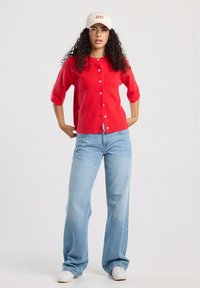 Nainen, jolla on kihara hius, pukeutuneena punaiseen napitettavaan villapaitaan, vaaleansinisiin leveälahkeisiin farkkuihin, valkoisiin tennareihin ja valkoiseen lippalakkiin, seisoo kädet lanteilla.