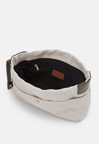 Jasnobeżowa torba z płótna z ciemną czarną podszewką, wyposażona w kieszeń na zamek, brązową skórzaną metkę oraz oliwkowozielony pasek. Zaokrąglony, miękki kształt.