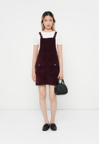 Robe salopette en velours côtelé bordeaux avec deux poches avant, bretelles épaisses et apparence dorée, associée à un petit sac à main noir et des chaussures noires.