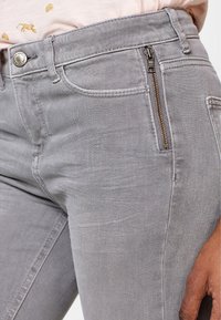 Jean en denim gris avec une légère texture, une poche zippée sur le côté et des poches avant classiques avec une fermeture à bouton métallique.
